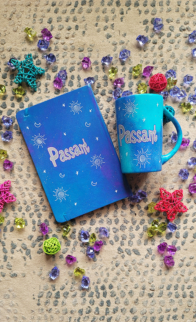 A Blue Galaxy hand painted mug and notebook with the name "Passant" with small moons stars and suns scattered all over.