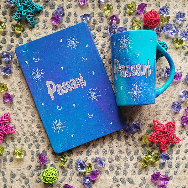 A Blue Galaxy hand painted mug and notebook with the name "Passant" with small moons stars and suns scattered all over.