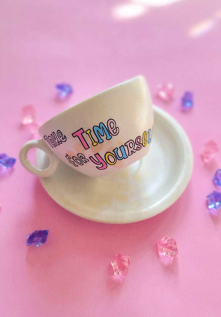 A Pearl white hand painted cup and saucer with gold geomteric patterns in the background and the words "Take time for Yourself"