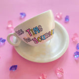 A Pearl white hand painted cup and saucer with gold geomteric patterns in the background and the words 