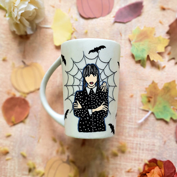 An Ivory hand painted mug with a gothic girl drawn in the center and a web behind her with bats all over the mug