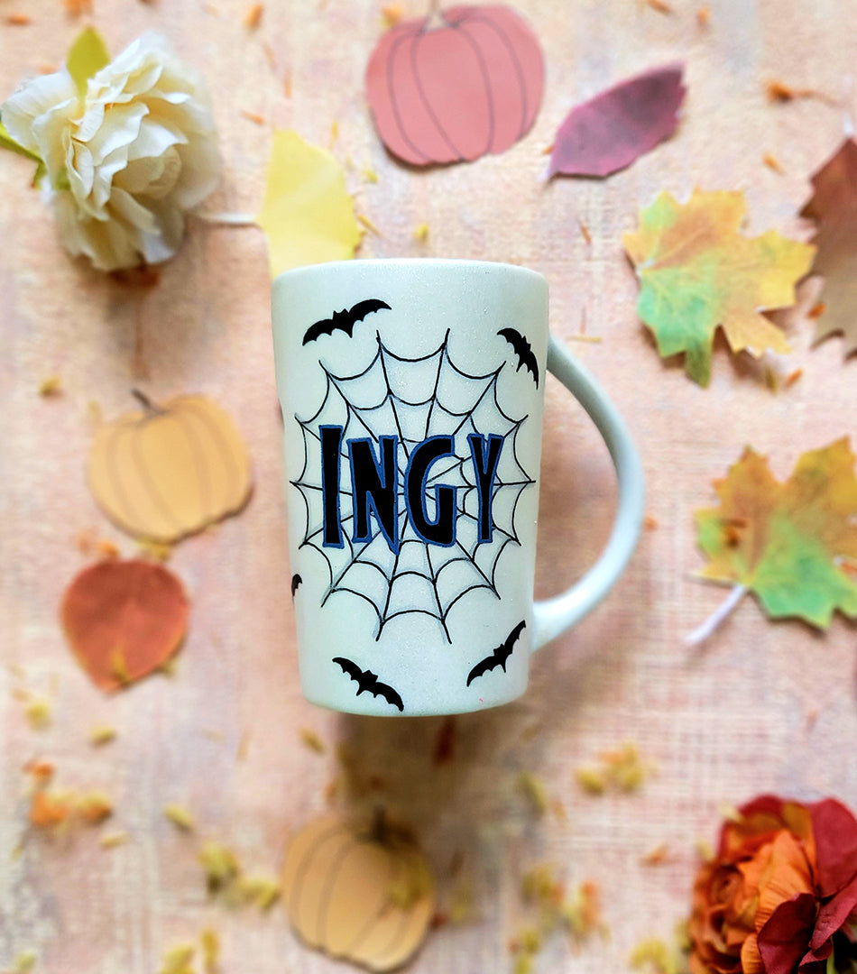 An Ivory hand painted mug with the name "Ingy " written on it and behind it there is a web, with bats scattered all over