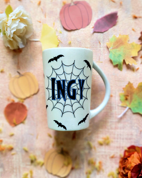 An Ivory hand painted mug with the name "Ingy " written on it and behind it there is a web, with bats scattered all over