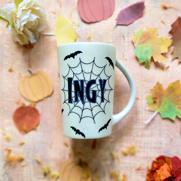 An Ivory hand painted mug with the name "Ingy " written on it and behind it there is a web, with bats scattered all over