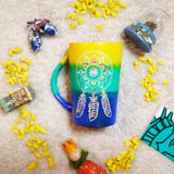 A gradient hand painted mug (yellow, green, blue) with a dreamcatcher in white and colored in pink, purple, orange, yellow, green and blue