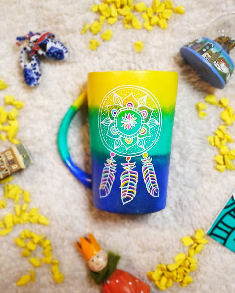 A gradient hand painted mug (yellow, green, blue) with a dreamcatcher in white and colored in pink, purple, orange, yellow, green and blue